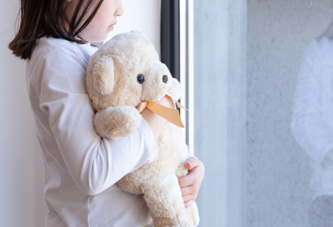 ぬいぐるみを抱えて窓の外を眺める子ども。